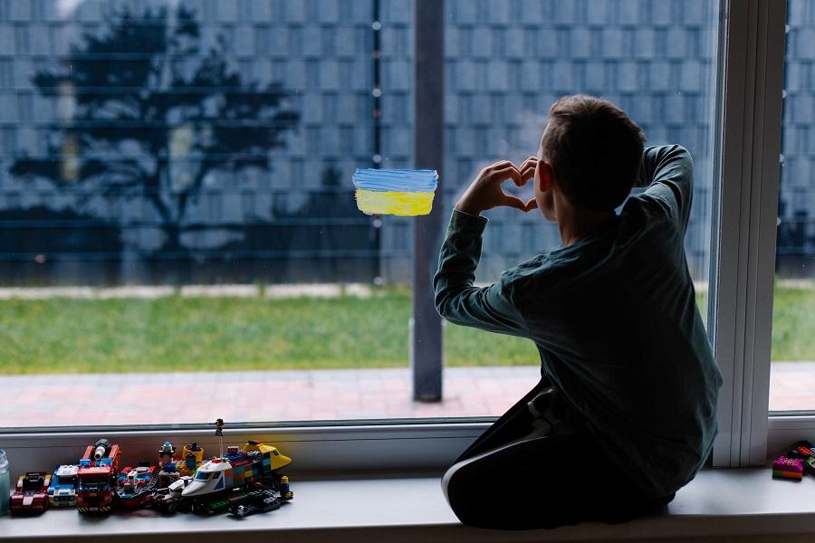 Photograph of a child seen from behind, looking through a glass window where the flag of Ukraine has been painted, while making a heart with his hands